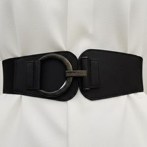 Puede incluir: Un cinturón elástico negro con una hebilla grande de color plateado. El cinturón tiene un diseño ancho y está hecho de cuero sintético. La hebilla tiene forma circular grande con un cierre rectangular. El cinturón se muestra sobre una prenda blanca.
