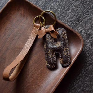 Op de afbeelding: Een bruine leren sleutelhanger met een gouden sleutelring en een bruine leren hanger in de vorm van de letter H. De letter H is versierd met een bruin en beige patroon.