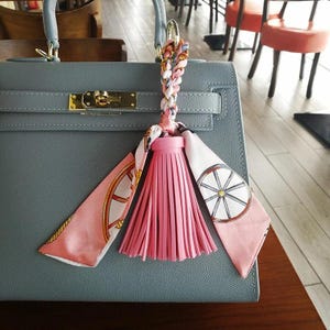 以下が含まれることがあります： グレーの革製のハンドバッグに、ピンクのタッセルキーホルダーが付いている。キーホルダーにはピンクのタッセルと、車輪のデザインが施された白とピンクのパターンのスカーフが付いている。