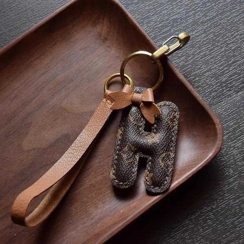 Op de afbeelding: Een bruine leren sleutelhanger met een gouden metalen ring en een bruine leren hanger in de vorm van de letter H. De hanger is versierd met een bruin en beige gepatenteerd stof.