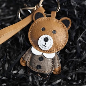 May include: A brown leather bear keyring with a gold-coloured ring and strap. The bear has a white face, black eyes, and a black nose. It is wearing a grey jacket with black buttons and a white collar.
