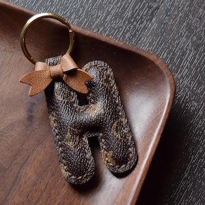 Op de afbeelding: Een bruine leren sleutelhanger met een monogrammotief en een bruine leren strik. De sleutelhanger is in de vorm van de letter H.