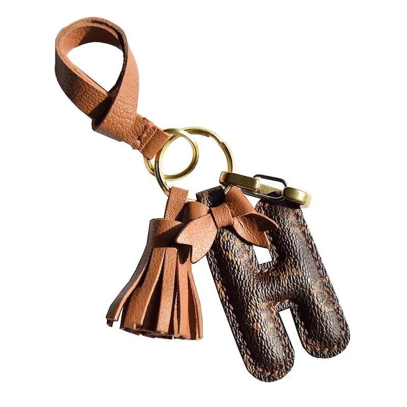 Op de afbeelding: Een bruine leren sleutelhanger met een bruine en beige monogram letter H bedel, een bruine leren kwast en een gouden sleutelring.