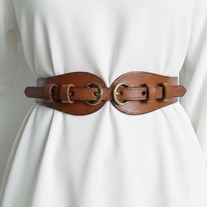 May include: A brown leather belt with a wide, curved design. The belt has two large gold-coloured metal rings at the centre and smaller gold-coloured metal rings on the sides. The belt is designed to cinch the waist and has a buckle closure.