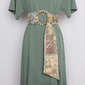 Könnte beinhalten: Ein salbeigrünes Kleid mit kurzen Ärmeln und einem dekorativen Gürtel. Der Gürtel hat eine goldfarbene ovale Schnalle mit Perlenakzenten und einen gemusterten Stoff mit beige, blauen und roten floralen Designs.