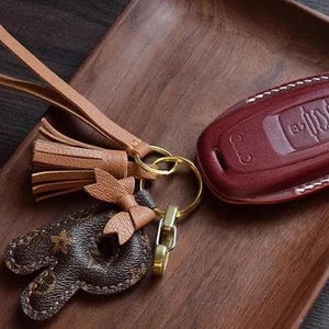 Op de afbeelding: Een bruine leren sleutelhanger met een kwast en een bruine leren sleutelhanger met een gegraveerde hanger. De sleutelhanger heeft een wit stikseldetail en een rood lederen interieur.