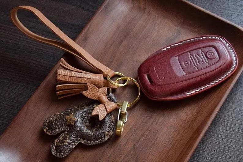 Op de afbeelding: Een bruine leren sleutelhanger met een kwast en een bruine leren sleutelhanger met een gegraveerde hanger. De sleutelhanger heeft een wit stikseldetail en een rood lederen interieur.