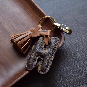 Op de afbeelding: Een bruine leren sleutelhanger met een kwast en een hanger in de vorm van de letter H. De hanger is gemaakt van bruin leer met een monogram-patroon.