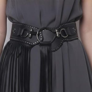 Może przedstawiać: Czarny pas z szerokim, ćwiekowanym wzorem i dużą metalową klamrą. Pas posiada frędzle po lewej stronie i szereg małych metalowych ćwieków wzdłuż krawędzi. Pas jest noszony na szarej sukience.