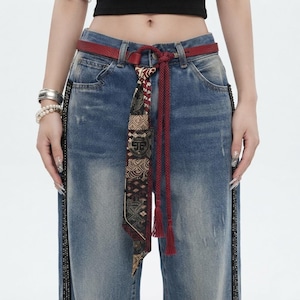 May include: Blue denim jeans featuring a dark wash with distressed detailing, side panels with metallic embellishments, and a red leather belt accented by a patterned fabric tie and a braided red cord with tassels hanging from the waist.