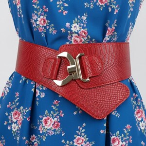 Puede incluir: Un cinturón rojo ancho con un patrón texturizado similar a la piel de serpiente y una hebilla dorada. El cinturón se usa sobre un vestido azul floral, creando una cintura definida y agregando un toque de color al atuendo.