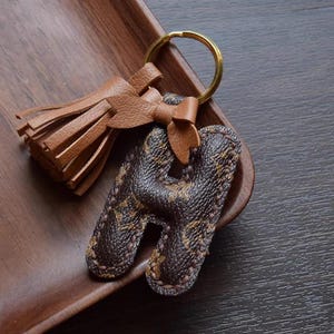Op de afbeelding: Een bruine leren sleutelhanger met een kwast en een letter H-bedel. De bedel is gemaakt van bruin leer met een monogram-patroon.