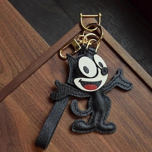 May include: Black leather keychain featuring a cartoon cat with white eyes and a red mouth. The keychain has a gold metal ring and a black leather strap.