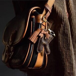 Op de afbeelding: Een bruine leren schoudertas met zwarte lederen afwerking en een bruine leren band. De tas heeft een sleutelhanger met een bruine leren kwast en een bruine leren hanger.