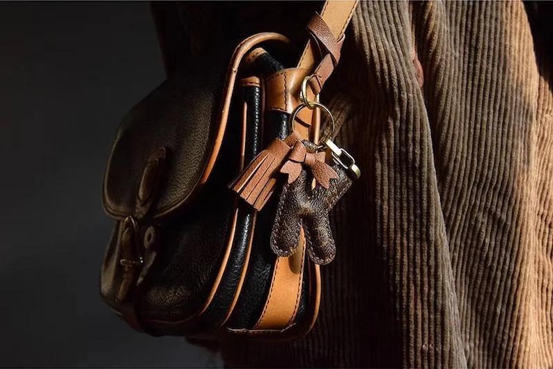 Op de afbeelding: Een bruine leren schoudertas met zwarte lederen afwerking en een bruine leren band. De tas heeft een sleutelhanger met een bruine leren kwast en een bruine leren hanger.