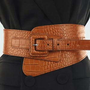 May include: A wide, tan-coloured belt featuring a textured, embossed crocodile skin pattern. The belt includes a large, matching tan square buckle with a silver-toned metal prong and a long strap that secures through the buckle for an adjustable fit.