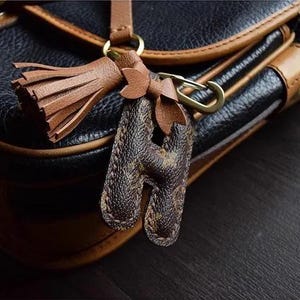 Op de afbeelding: Een bruine leren tas met een gouden metalen sluiting en een bruine leren kwast sleutelhanger met een monogram letter H.