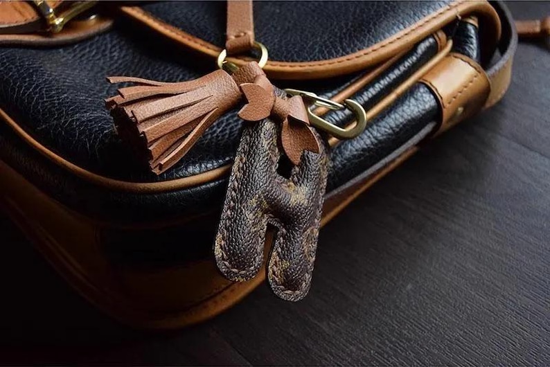 Op de afbeelding: Een bruine leren tas met een gouden metalen sluiting en een bruine leren kwast sleutelhanger met een monogram letter H.