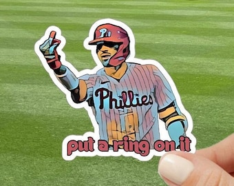 May include: A cartoon illustration of a baseball player wearing a red and blue Phillies jersey with the text "Phillies" and "put a ring on it".