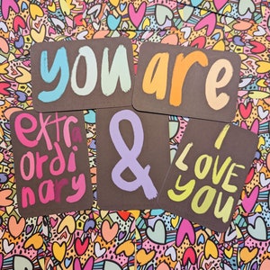 May include: Five cards with colorful, handwritten messages on a black background. The messages are "you are", "extraordinary", "&", "I love", and "you". The cards are on a background of colorful hearts and swirls.