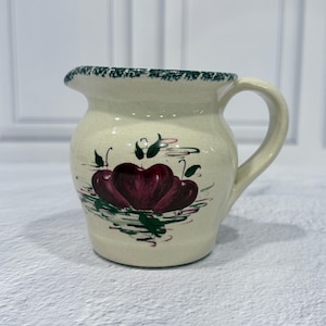以下が含まれることがあります： クリーム色のセラミックピッチャー。緑色の縁とハンドルが付いています。ピッチャーには、緑の葉と茎が付いた、濃い赤色のリンゴ3つの手描きのデザインが施されています。装飾的なキッチン用品です。