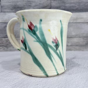 Op de afbeelding: Crèmekleurige keramische kan met een handvat, versierd met handgeschilderde groene stelen, rode bloemknoppen en gele accenten. De kan heeft een licht uitlopende rand en een gladde, glanzende afwerking. Een uniek stuk voor het serveren van dranken.