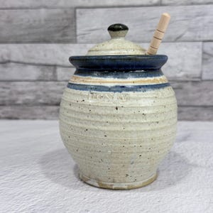 Puede incluir: Un tarro de miel de cerámica con tapa y cuchara de miel de madera. El tarro es de color blanquecino con rayas azules y beige y la palabra "HONEY" en relieve en la parte delantera. La tapa tiene un borde azul oscuro y un pequeño pomo negro.