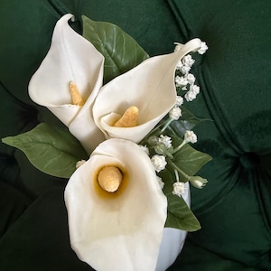 Puede incluir: Un arreglo floral que presenta tres calas blancas con espádices amarillos prominentes, acentuado por pequeñas flores blancas de paniculata y varias hojas de color verde oscuro, todo reunido en un recipiente blanco.