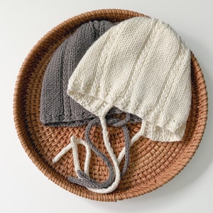 Puede incluir: Dos gorros de bebé tejidos, uno gris y uno blanco, con lazos, en una cesta tejida.