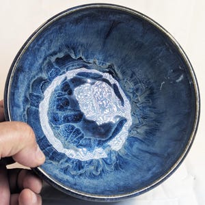 May include: A round ceramic bowl featuring a deep blue glaze with swirling, marbled patterns in lighter shades of blue and white. The interior surface displays a glossy finish with a distinct, textured appearance, and the rim is dark.