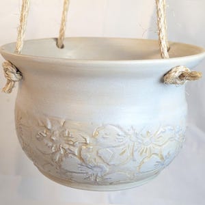 May include: A light gray ceramic hanging planter with a wide, flared rim and a rounded body. The lower portion features a raised, embossed floral pattern. The planter is suspended by thick, natural-colored jute rope knotted at the sides.