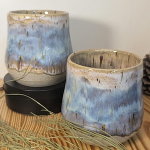 May include: Two ceramic tumblers with a blue and tan glaze. The handmade cups have a rounded shape and a smooth, glossy finish. The blue glaze has a watercolor effect, with streaks of tan and brown. The cups are on a wooden surface.