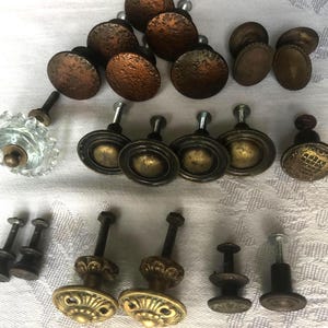 May include: A collection of antique cabinet knobs in various styles and finishes. The knobs include textured bronze, brass, and clear glass options. Some knobs have a round, flat design, while others feature ornate detailing.