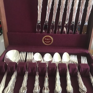 May include: A set of silverware in a wooden box with a burgundy velvet lining. The silverware is made of silver and has an ornate design. The box has a label that says "Naken International Silverware Chest".