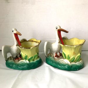 May include: A pair of vintage ceramic pelican figurines. Each pelican has a white body, red beak, and is perched next to a yellow flower-shaped vase with green leaves. The base is a green, wave-like design.