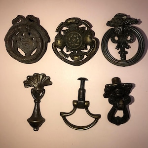 May include: Six antique bronze drawer pulls and knobs. The pulls feature ornate designs, including a ring pull, a fan shape, and floral motifs. The hardware is in various shapes and sizes, with a vintage aesthetic.