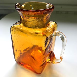 May include: Amber-colored glass pitcher with a square body and a clear, curved handle. The pitcher has a textured surface and a flared rim. A decorative piece for serving beverages.