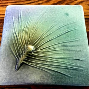 May include: Square ceramic coaster with a detailed embossed pine needle design. The coaster features a gradient of colors, transitioning from blue to green, with a central light yellow accent. The coaster is a decorative and functional item.