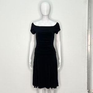 Puede incluir: Un vestido negro con escote de hombros descubiertos y mangas cortas. El corpiño está fruncido y la falda se ensancha desde la cintura. El vestido se exhibe en un maniquí blanco sobre un fondo blanco.