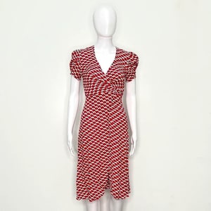 May include: A knee-length dress featuring a V-neckline and short puff sleeves. The garment is patterned with a repeating red and white geometric design. It has a wrap-style bodice that gathers at the waist, flowing into a straight skirt silhouette.