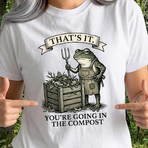 Op de afbeelding: Wit T-shirt met een cartoon kikker die een tuinvork vasthoudt, naast een houten compostbak gevuld met bladeren. De tekst luidt "THAT'S IT. YOU'RE GOING IN THE COMPOST."