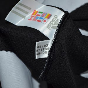 May include: Close-up of a black garment with a white Adidas label. The label shows the Adidas logo, various flags, and a smaller label with the numbers "06/03", "021786", and "AHJ001". The garment has a white stripe.