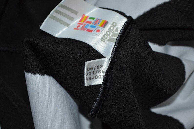May include: Close-up of a black garment with a white Adidas label. The label shows the Adidas logo, various flags, and a smaller label with the numbers "06/03", "021786", and "AHJ001". The garment has a white stripe.