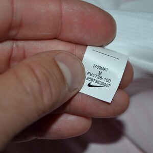 May include: A white fabric garment tag featuring the black Nike swoosh logo, the size M, and the printed text 2403MAT, FV1738-100, and 196975639007. The tag is rectangular with a dashed line at the top and is attached to a white textile material.