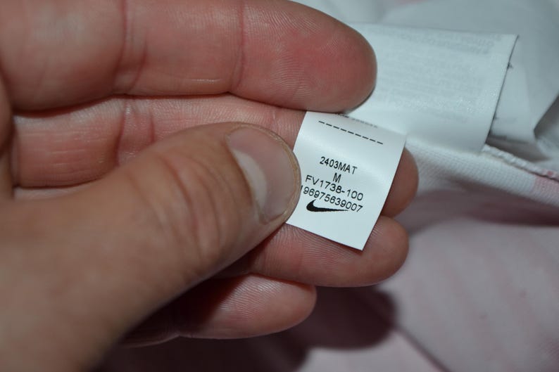 May include: A white fabric garment tag featuring the black Nike swoosh logo, the size M, and the printed text 2403MAT, FV1738-100, and 196975639007. The tag is rectangular with a dashed line at the top and is attached to a white textile material.