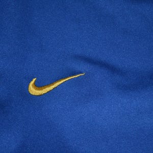 May include: A close-up of a royal blue athletic top featuring a gold Nike swoosh logo. The fabric has a textured finish. The top is likely made of a breathable material, suitable for sports.