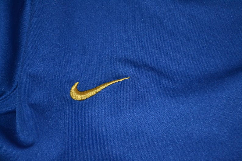 May include: A close-up of a royal blue athletic top featuring a gold Nike swoosh logo. The fabric has a textured finish. The top is likely made of a breathable material, suitable for sports.