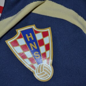 May include: Close-up of a navy blue fabric with a Croatian soccer team emblem. The emblem is a shield shape with a red and white checkered pattern, blue letters, and a soccer ball at the bottom. A yellow stripe and red and white checkered fabric are also visible.