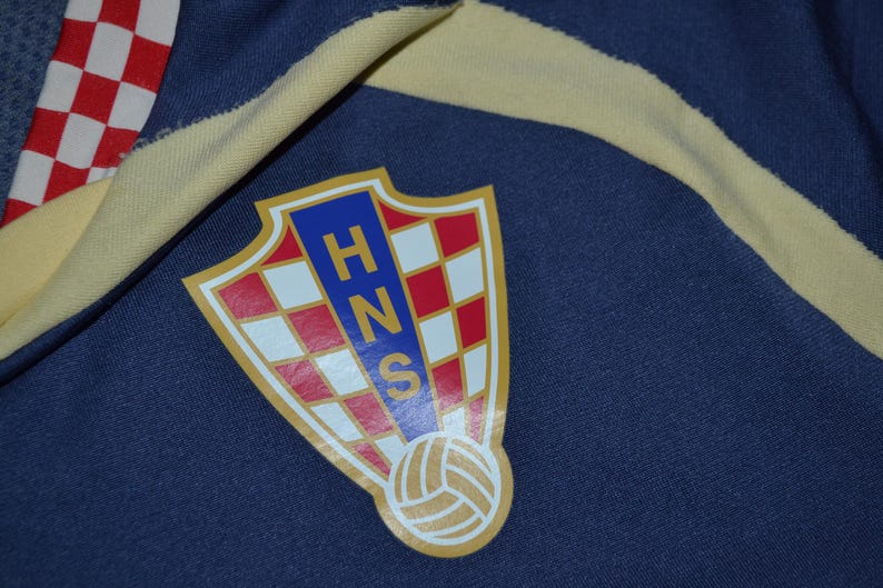 May include: Close-up of a navy blue fabric with a Croatian soccer team emblem. The emblem is a shield shape with a red and white checkered pattern, blue letters, and a soccer ball at the bottom. A yellow stripe and red and white checkered fabric are also visible.