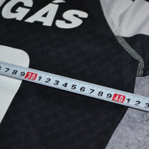 Puede incluir: Una camiseta gris oscuro con el texto blanco "QUIG&Aacute;S". Una regla muestra una medida de 30 a 40 cm. La camiseta tiene mangas a rayas blancas y grises. La tela parece ser un material de punto suave.